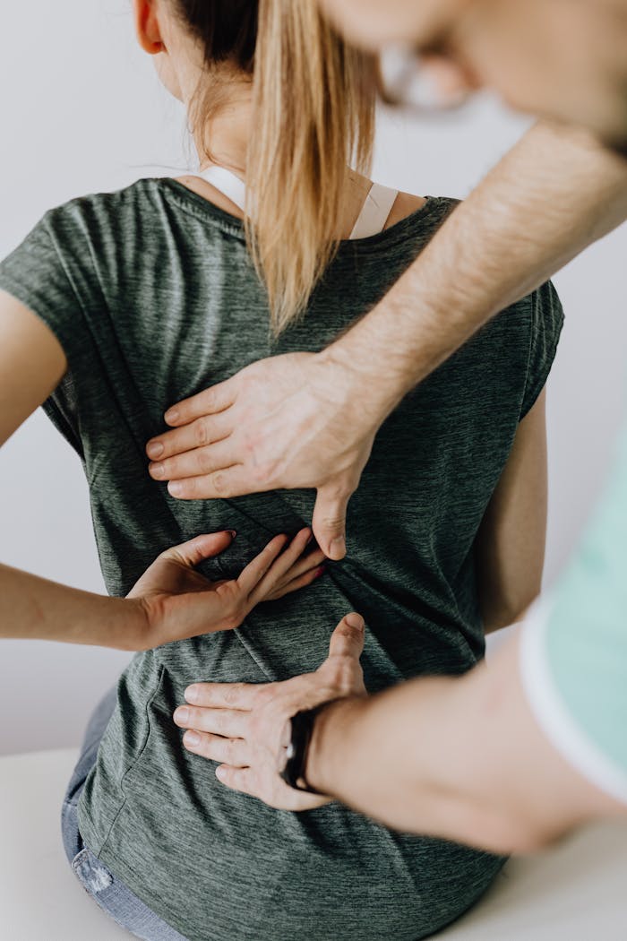 Top view of crop unrecognizable woman in casual wear showing chiropractor in eyeglasses and uniform pain spot on back while sitting on examination couch in hospital